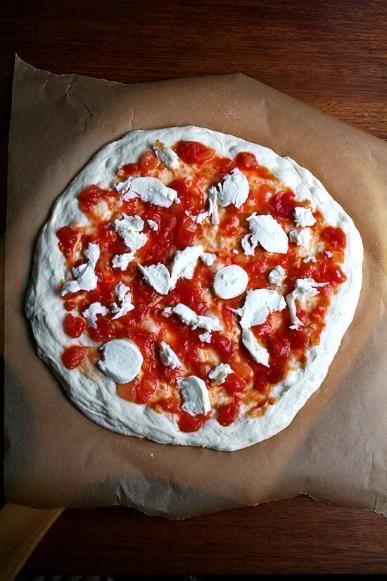 Stretched round of Lahey pizza dough on a pizza peel topped with sauce and cheese.