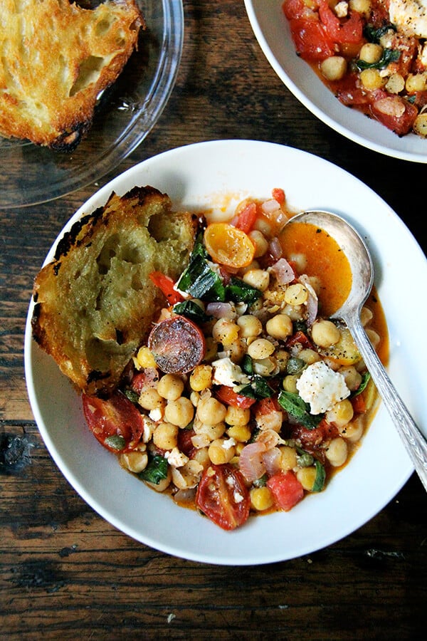 A bowl of stewy chickpeas with tomatoes, basil, feta, and olive oil toasted bread.
