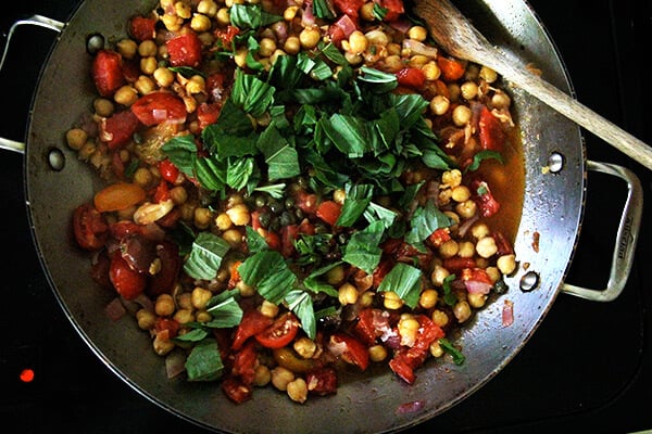 A sauté pan with chickpeas, olive oil, garlic, basil, and capers.