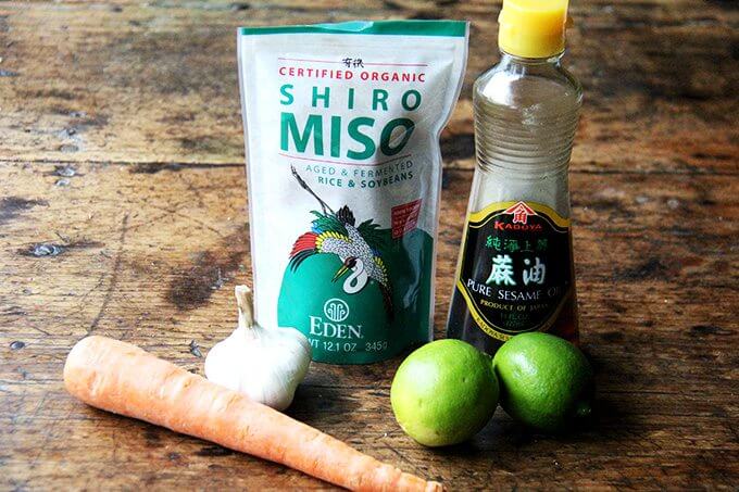 The ingredients for the dressing on a table: carrot, miso, garlic, limes, and sesame oil.