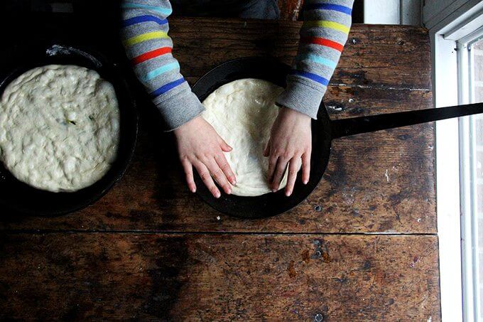 Making these skillet pizzas, I've come to realize, is a relatively good activity for involving children. For one, the dough doesn’t contain too many ingredients, and it’s forgiving—if flour spills out of the bowl, it’s easy to add more. More to the point, there are no eggs! Also, the many and varied steps of pizza making—mixing the dough, shredding the cheese, stretching and saucing the dough, topping the rounds—keeps the kids engaged. // alexandracooks.com A child spreads pizza dough in a skillet.