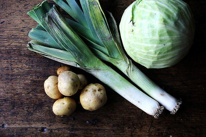 A board with potatoes, leeks, and a head of cabbage.