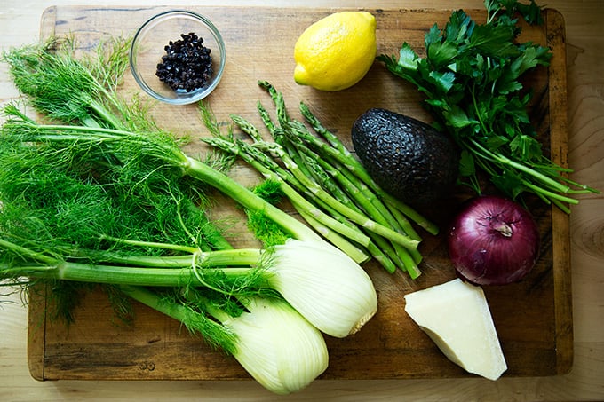 A cutting board holding the ingredients for the shaved fennel salad: fennel, asparagus, onion, lemon, parsley, parm, and currants.