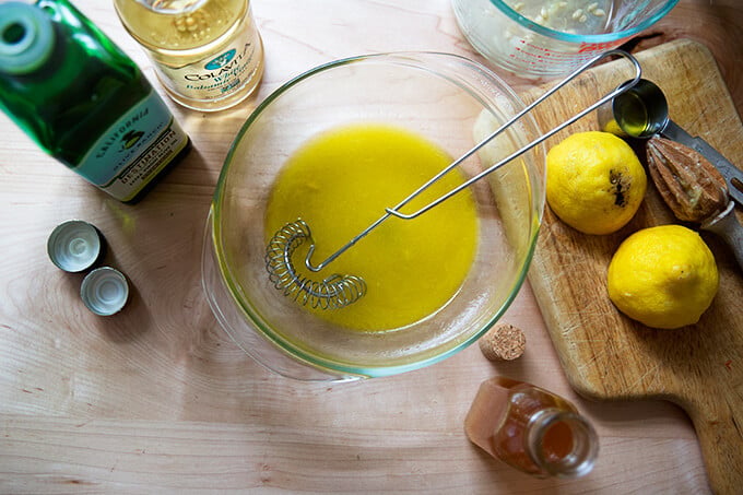 A bowl with a whisk and salad dressing aside bottles of olive oil, white balsamic, honey, and a halved lemon.