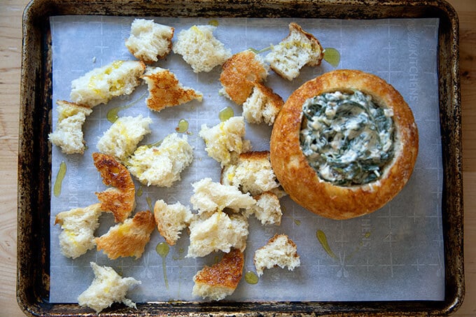 A sheet pan filled with a peasant bread bread bowl filled with spinach-artichoke dip.