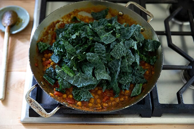 A skillet on the stovetop holding stewy chickpeas and tomatoes with a heap of kale on top.