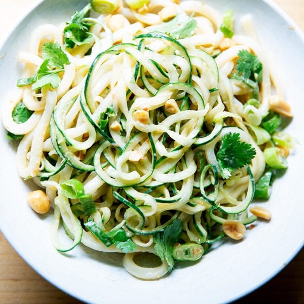 A bowl of peanut noodles with cucumbers, scallions and peanuts.
