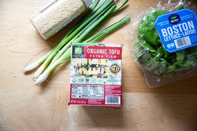 A block of extra-firm tofu on a counter aside a head of Boston lettuce, scallions, and sesame seeds.