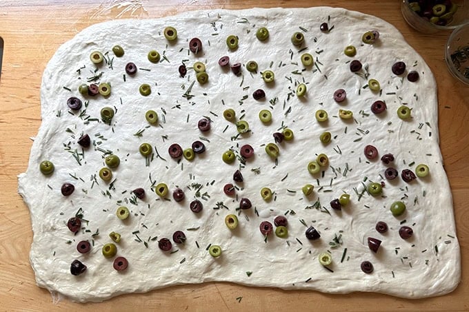 Sourdough dough stretched thinly across a work surface with olives and rosemary sprinkled across it.