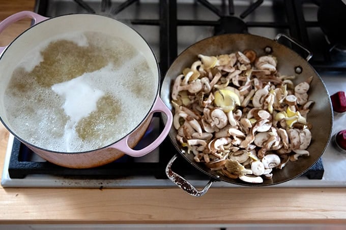 A large pot on the stovetop boiling pasta aside a large sauté pan filled with mushrooms.