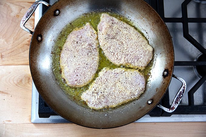 Three chicken cutlets frying in a large skillet on the stovetop.