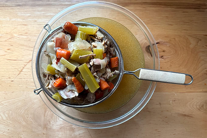 A strainer filled with boiled vegetables and chicken bones over a bowl of broth.