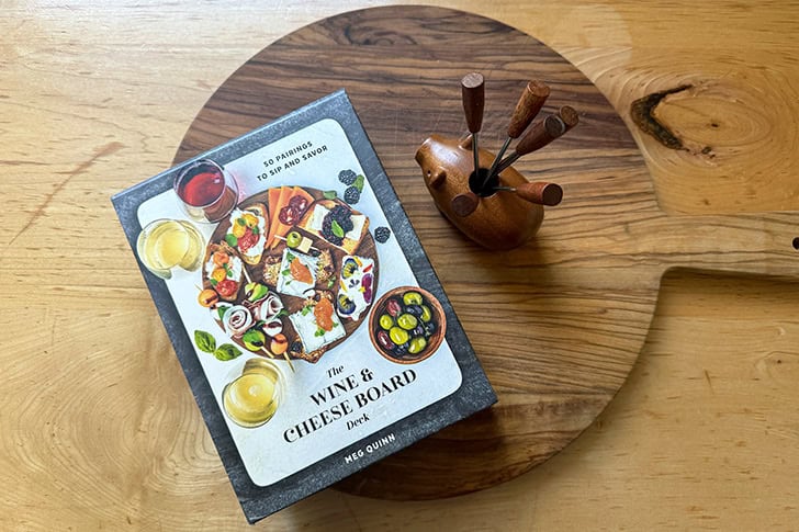 A cheeseboard with a wooden cheese pick pig on top as well as The Wine & Cheese Board Deck.