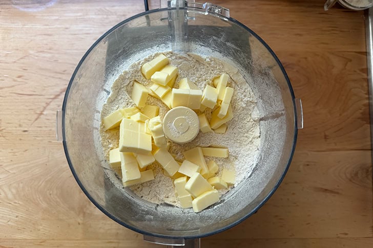 The butter to make cheater's croissant dough added to the bowl of a food processor holding the blended together dry ingredients.
